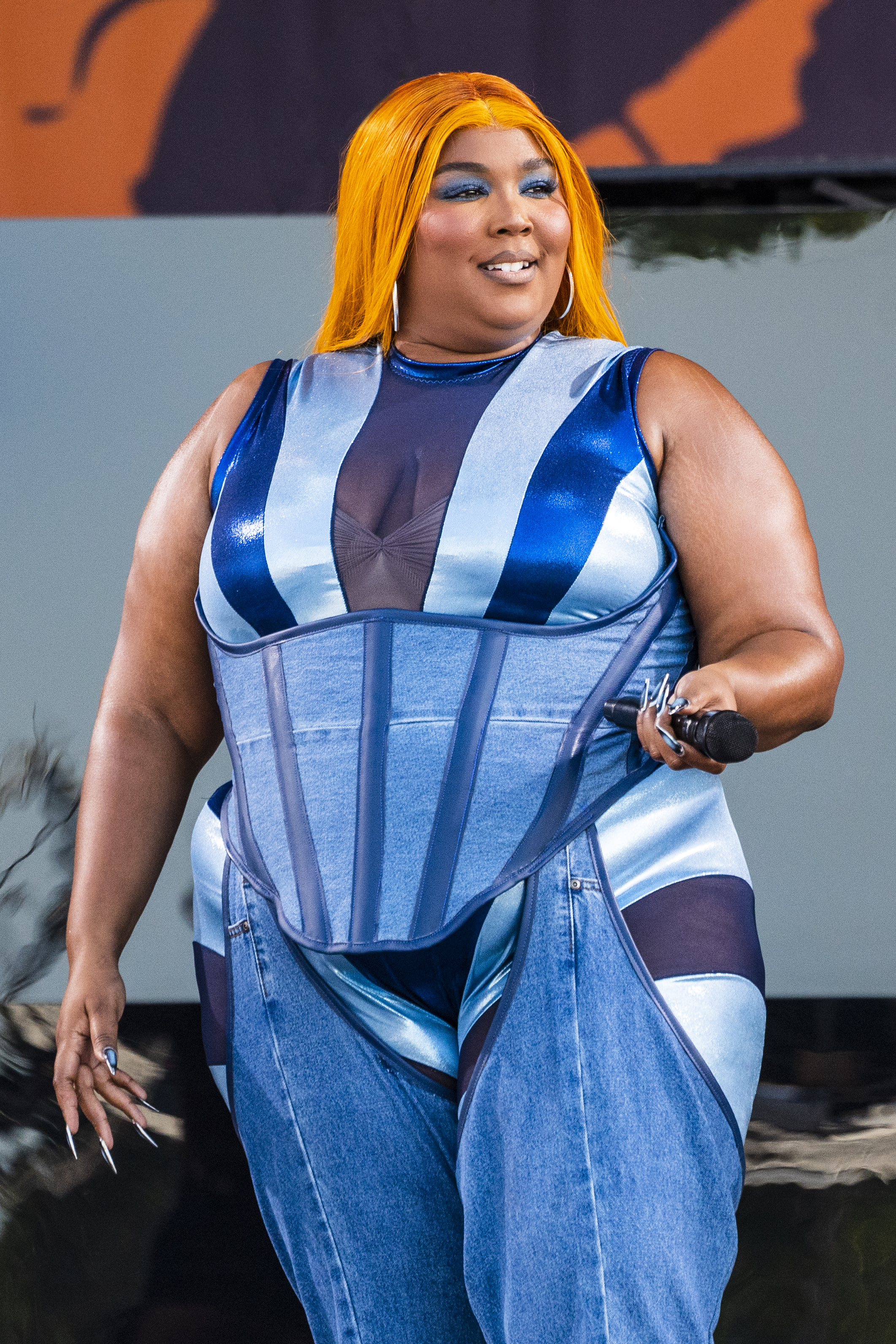 Lizzo performs on stage, wearing a denim jumpsuit with structured corset details and holding a microphone