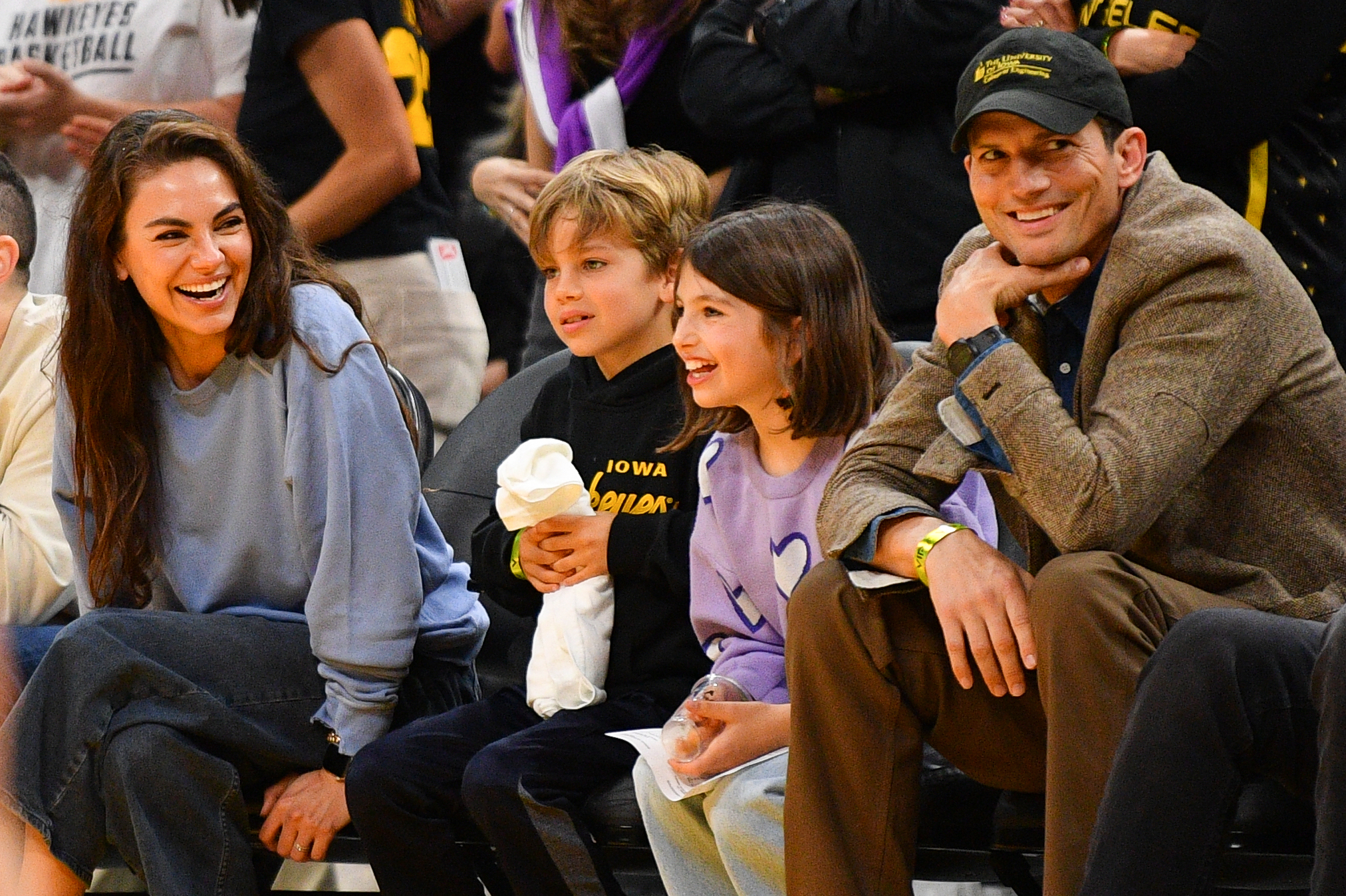 Mila Kunis and Ashton Kutcher attend a basketball game with their children. They are seated and smiling, enjoying the event