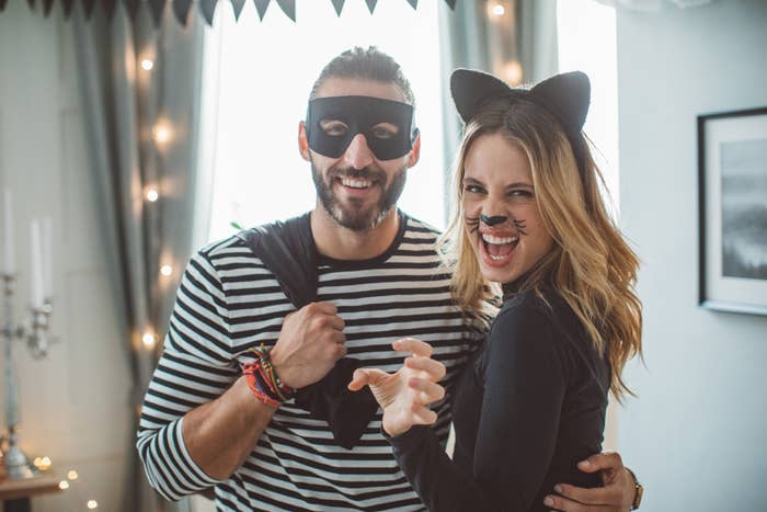 A man and woman in Halloween costumes. The man wears a striped shirt with a black mask, while the woman has cat ears, whiskers, and paws. Both are smiling