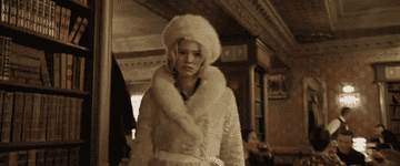 A woman in a luxurious fur coat and matching hat walks confidently through a richly decorated room and pulls out a gun