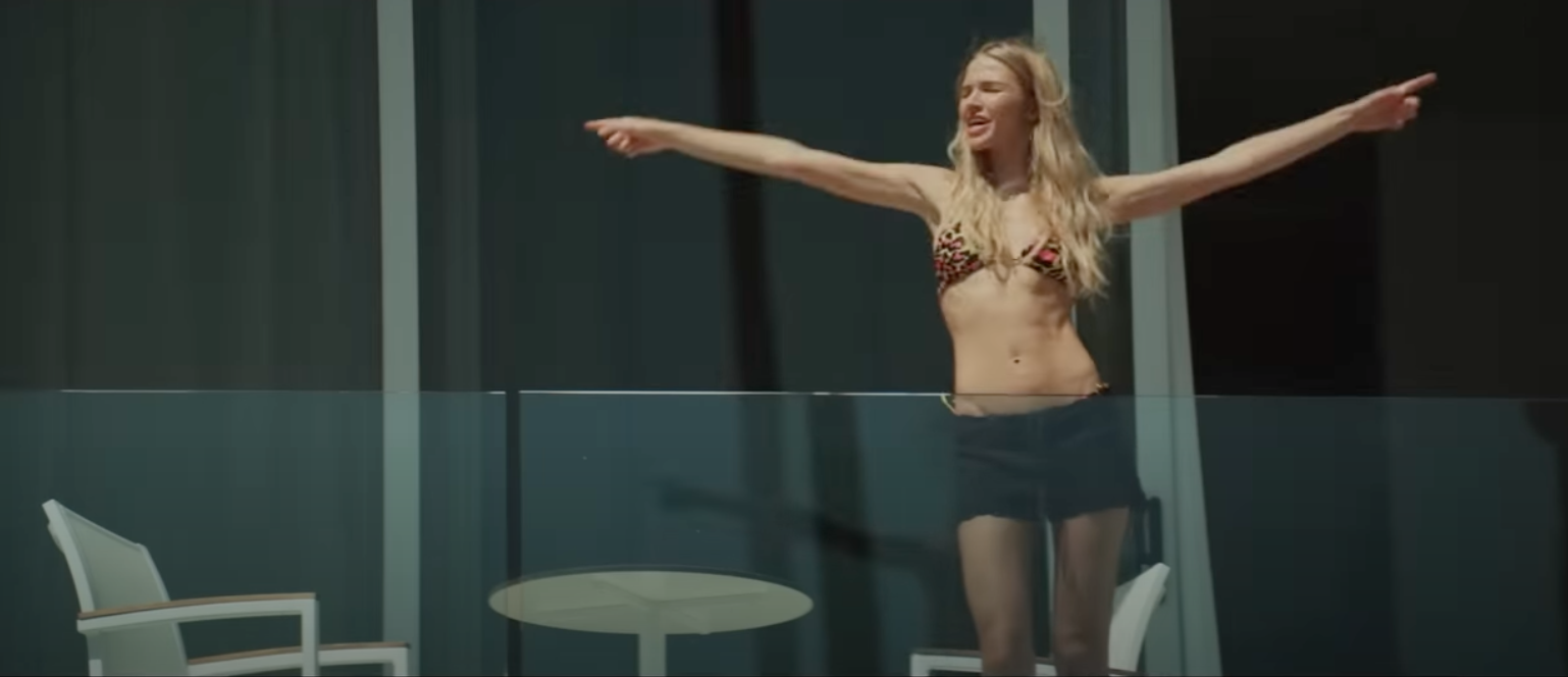 Sasha Luss in Sheroes, standing on a balcony in a bikini top and black shorts, with her arms extended outward