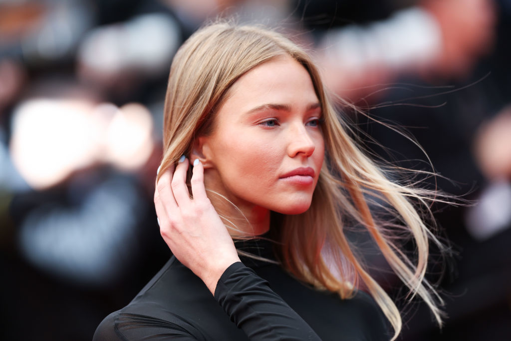 Sasha Luss in a sleek black outfit poses with one hand near her face