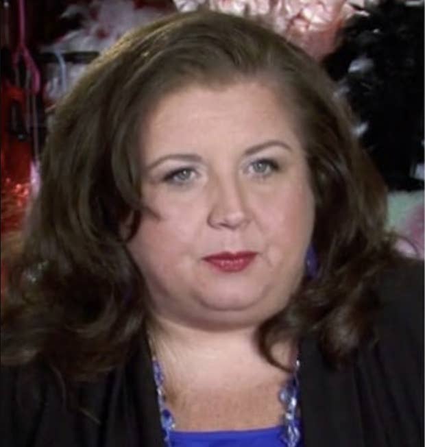 Abby Lee Miller, wearing a black jacket and blue top, poses for a photo with a neutral expression