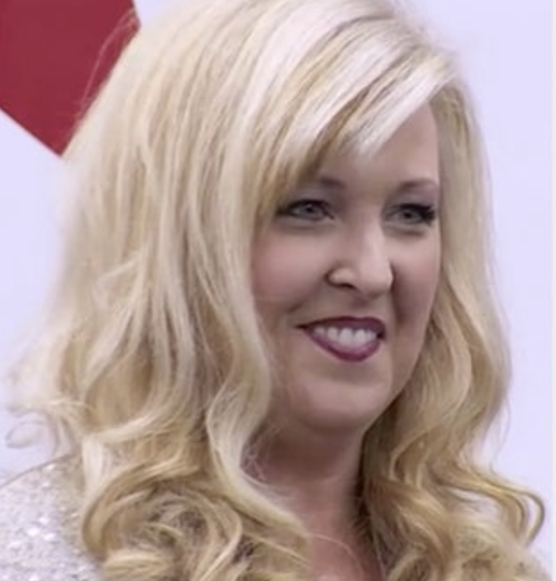 A woman with long blonde hair smiles while wearing a shiny top