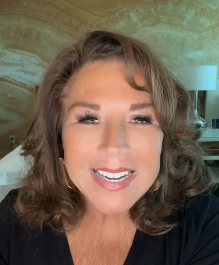 Close-up of Abby Lee Miller smiling, wearing a black top with curly, shoulder-length hair. She is in a room with a patterned wall and a bed visible in the background