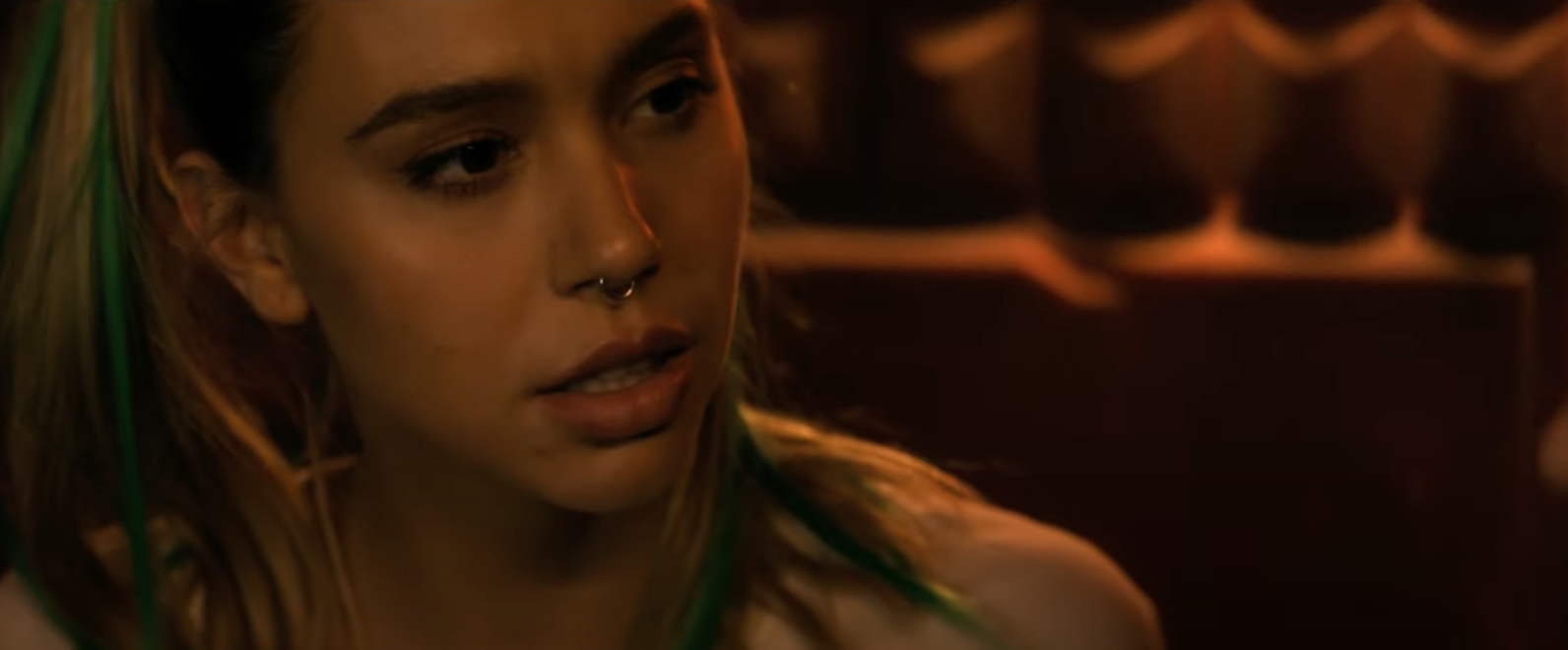 Alexis Ren in latency. her hair dyed is with green streaks, she is wearing a nose ring, and looks pensive while sitting indoors with dim lighting
