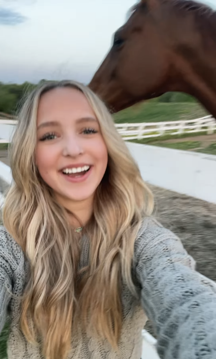 A person smiling in a selfie with a horse in the background, both in an outdoor setting near white fences