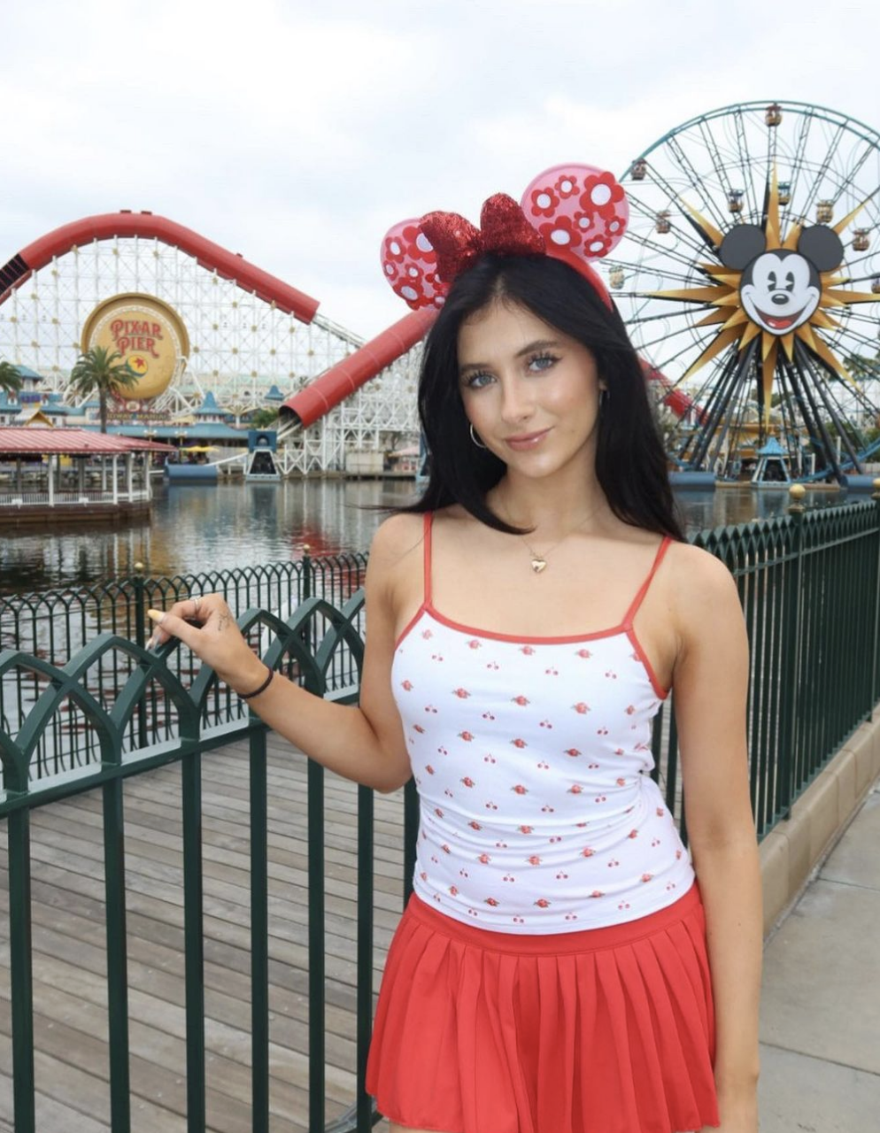 A woman wearing Minnie Mouse ears, a white tank top with small red patterns, and a red skirt poses in front of a theme park featuring a Ferris wheel with Mickey Mouse and a roller coaster