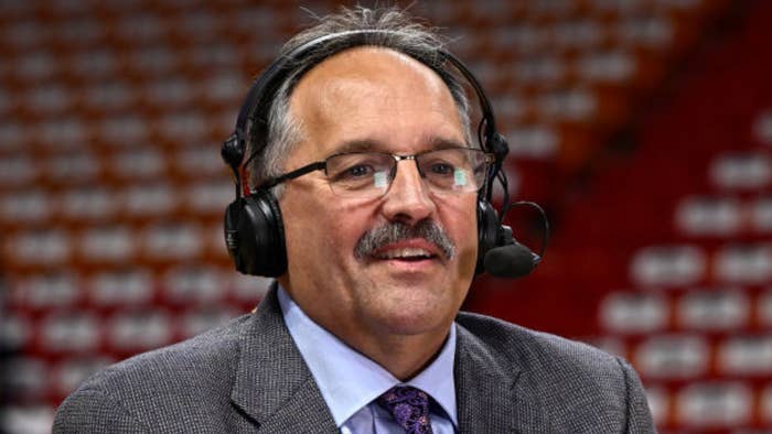 A man with glasses, a mustache, and wearing a headset, dressed in a gray suit jacket and tie, appears to be speaking or commentating
