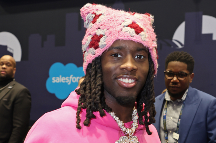 Kai Cenat wearing a pink knit hat and hoodie adorned with a large necklace smiles at a Salesforce event. Two people are in the background