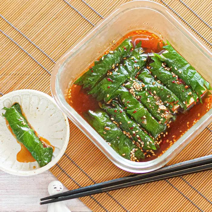 葉に包まれた料理がガラス容器に入っており、白い皿に一つ盛り付けられている。黒い箸と箸置きが見える。