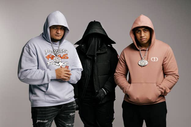 Three musicians, all wearing hoodies and chains, pose together. The person in the middle is dressed in black, with their face concealed by a hood