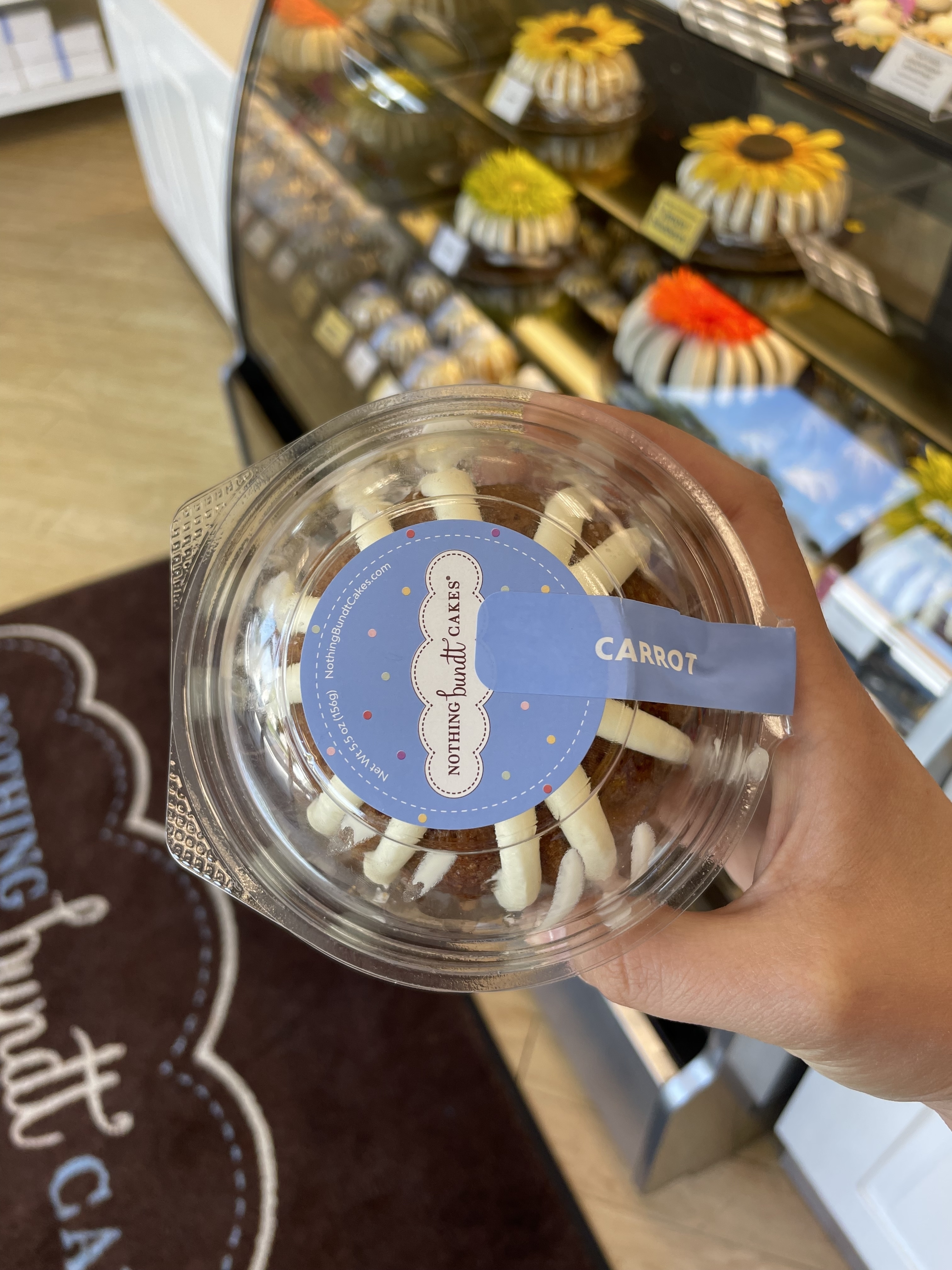 A hand holding a packaged carrot cake from "Nothing Bundt Cakes." A display case with various bundt cakes can be seen in the background