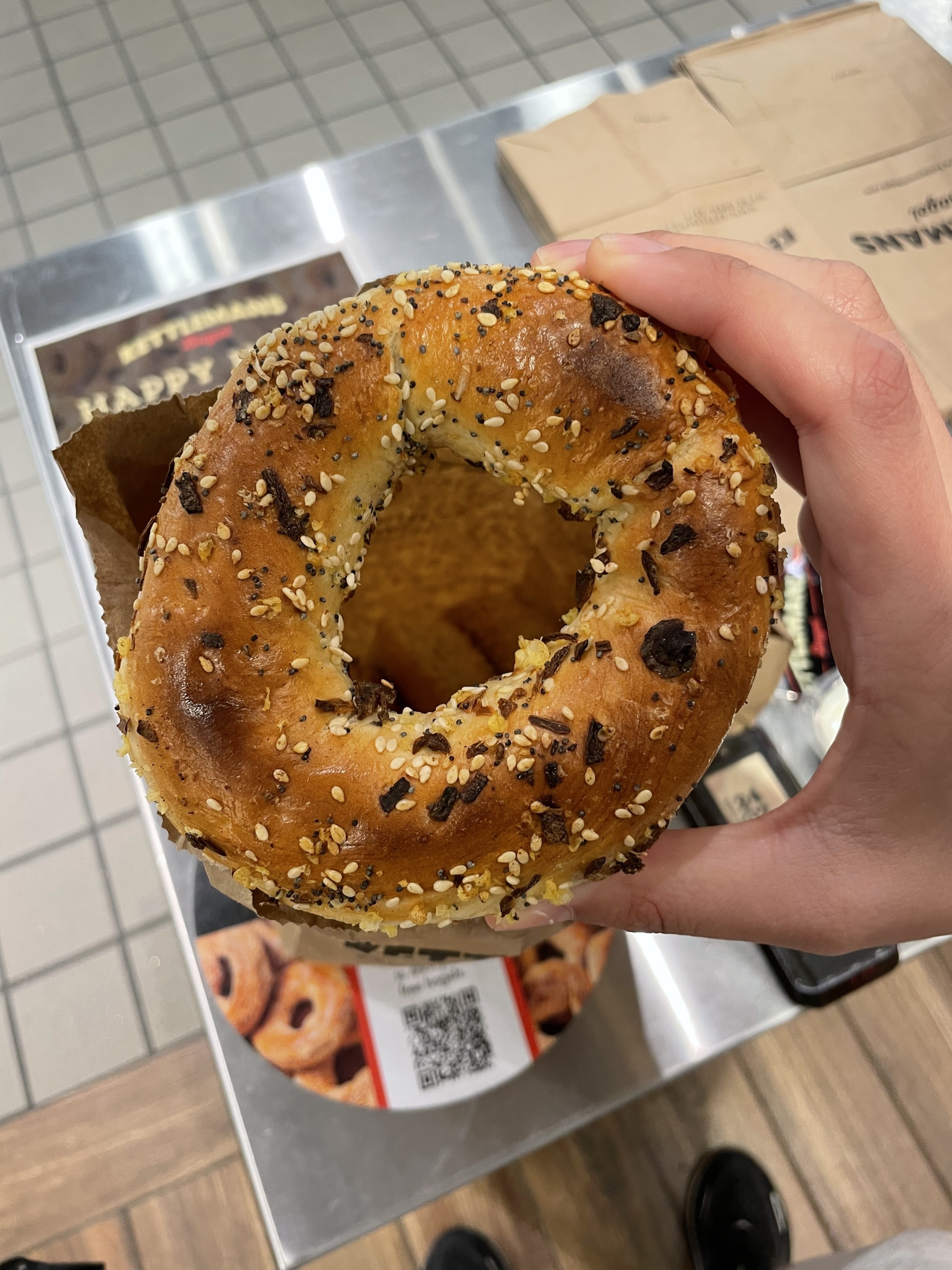 A hand holding an everything bagel over packaging on a table. The bagel has various seeds and seasoning on top