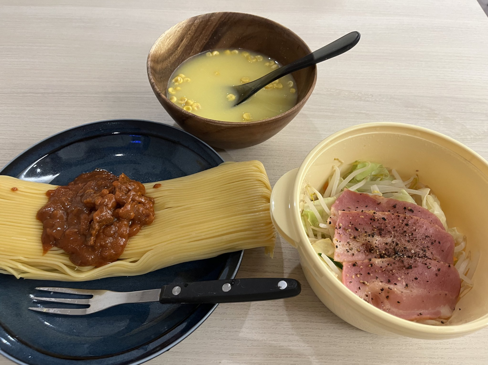 麺とミートソースが並んだ皿、スープの入った木のボウル、ハムと野菜が盛られたボウルとフォークが並んでいる。