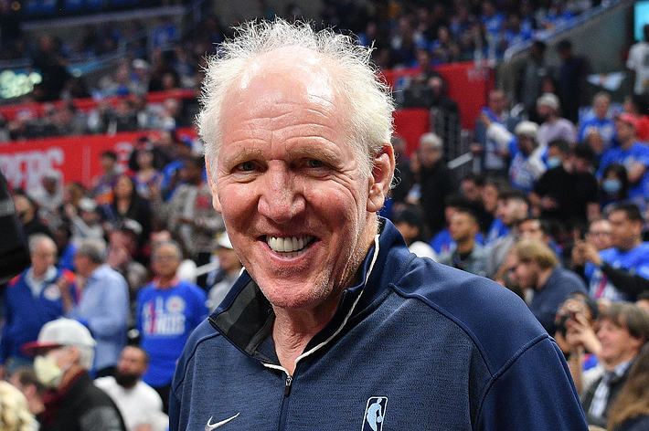 Bill Walton smiling in an arena filled with fans. He is wearing a dark Nike jacket with a basketball logo