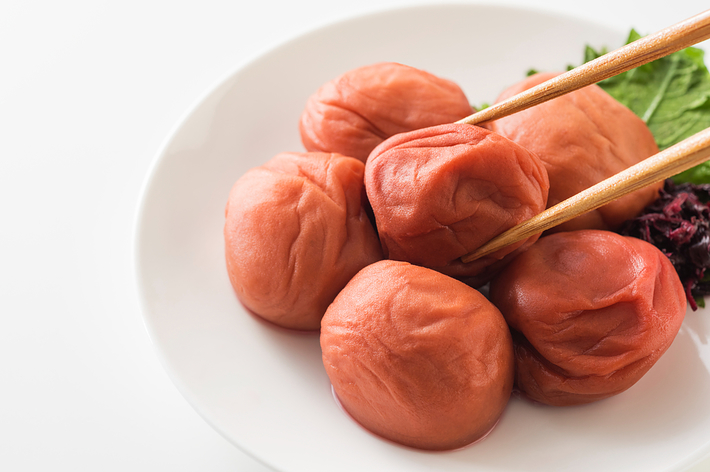 梅干しが白い皿に盛られており、箸で1つつまんでいる様子。背景には緑色の葉も添えられている。