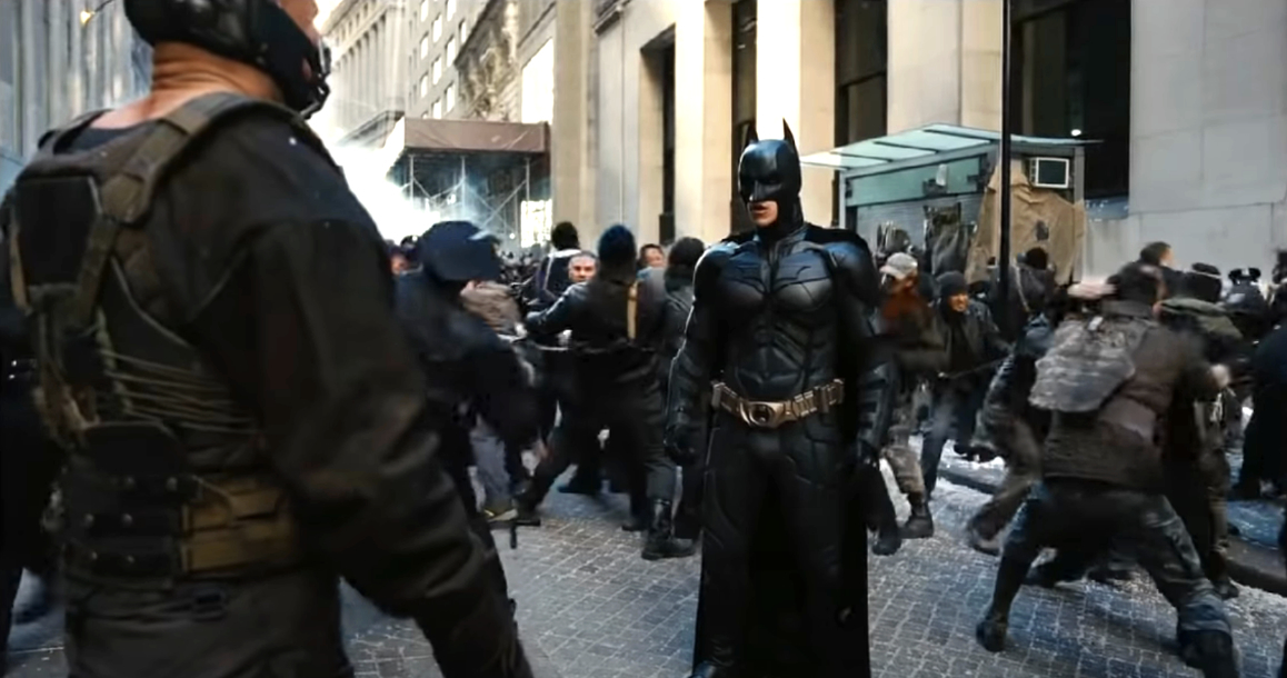 Batman, in his iconic suit, stands facing off against Bane amidst a chaotic street scene with fighters engaged in a battle