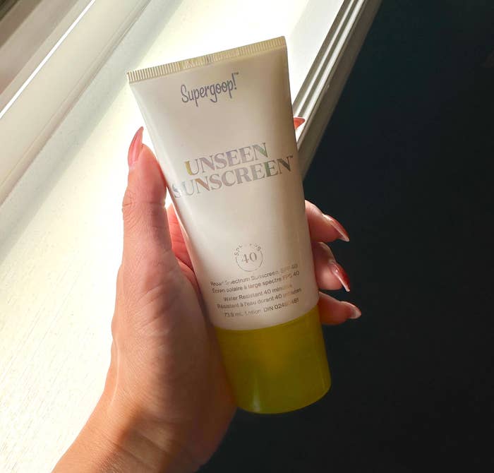 Hand holding a tube of Supergoop! Unseen Sunscreen SPF 40 near a window. The person is wearing a white watch and has polished nails
