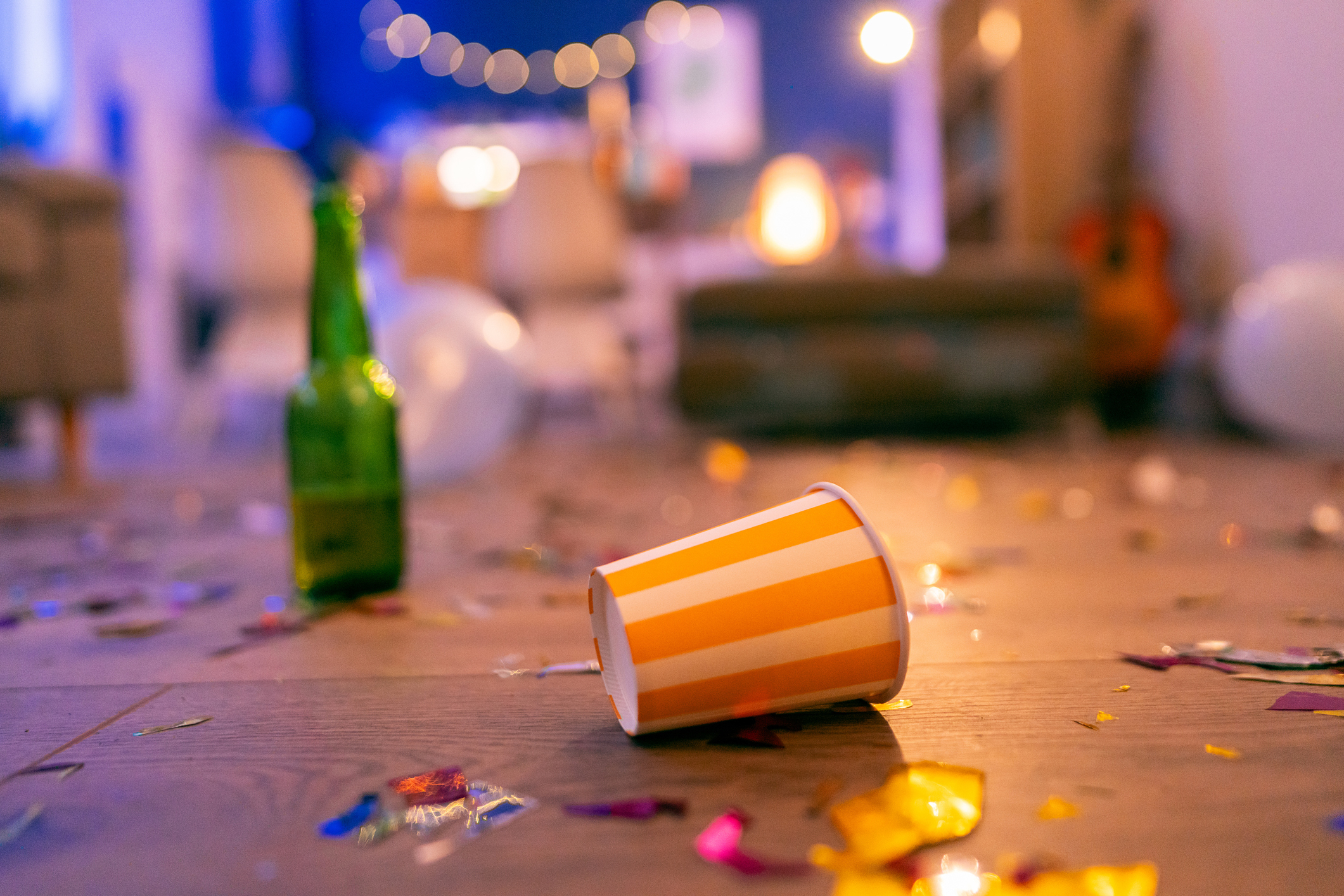 A messy room with an empty orange and white striped cup on the floor among confetti and a green bottle, likely after a party