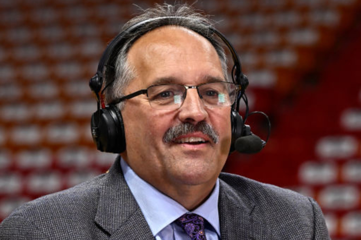Man with mustache, glasses, and headset speaks during a broadcast in an arena with empty seats in the background