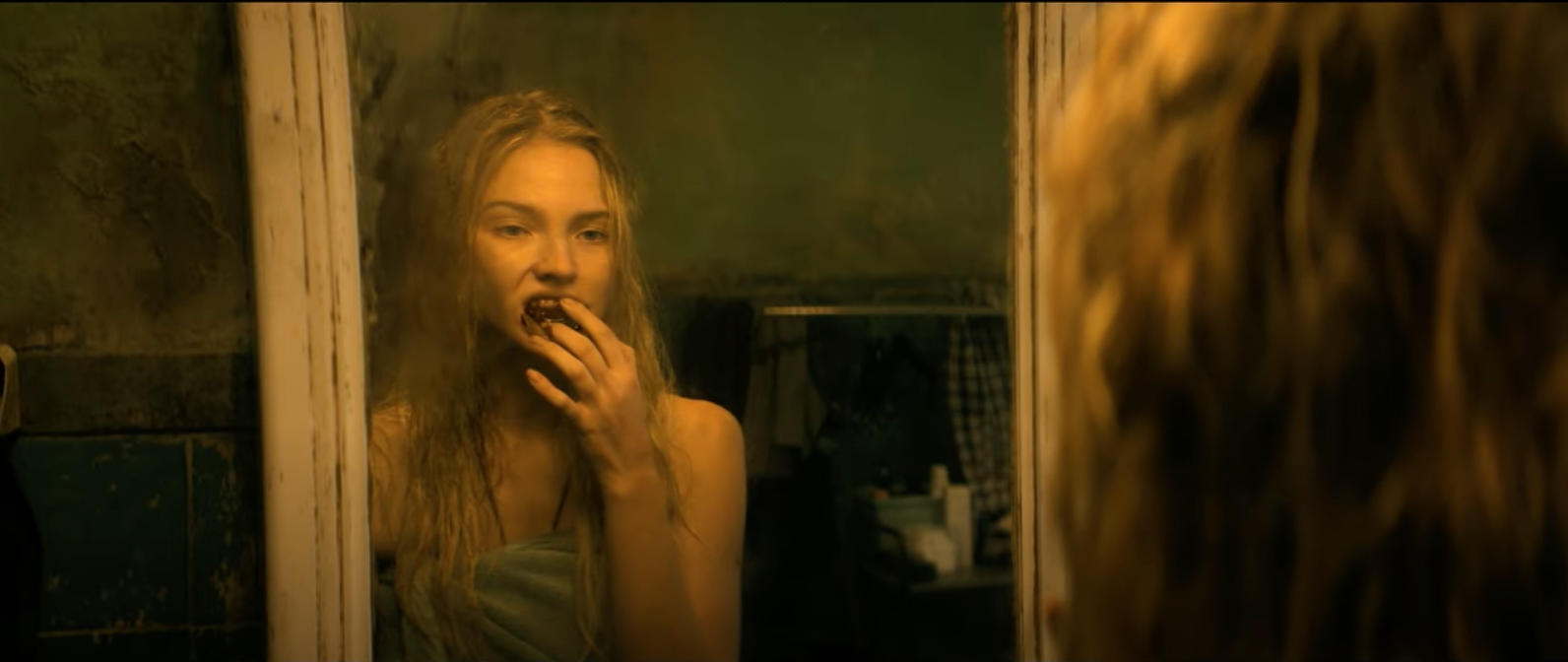 Sasha Luss, wrapped in a towel, gazes into a grimy mirror and touches her bloody teeth