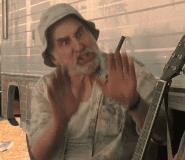 Bearded man emphatically gesturing with his hands while sitting next to a guitar. He looks frustrated and is wearing a bucket hat and a floral shirt