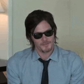 Norman Reedus wearing sunglasses and a tie, nodding while seated indoors