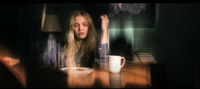 Sasha Luss in latency sits at a table with a mug and a plate. She appears tired or distressed