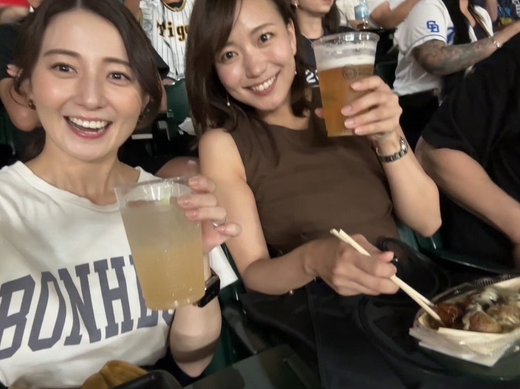 2人の女性が飲み物を手に持ちながら、観客席で笑顔を見せています。