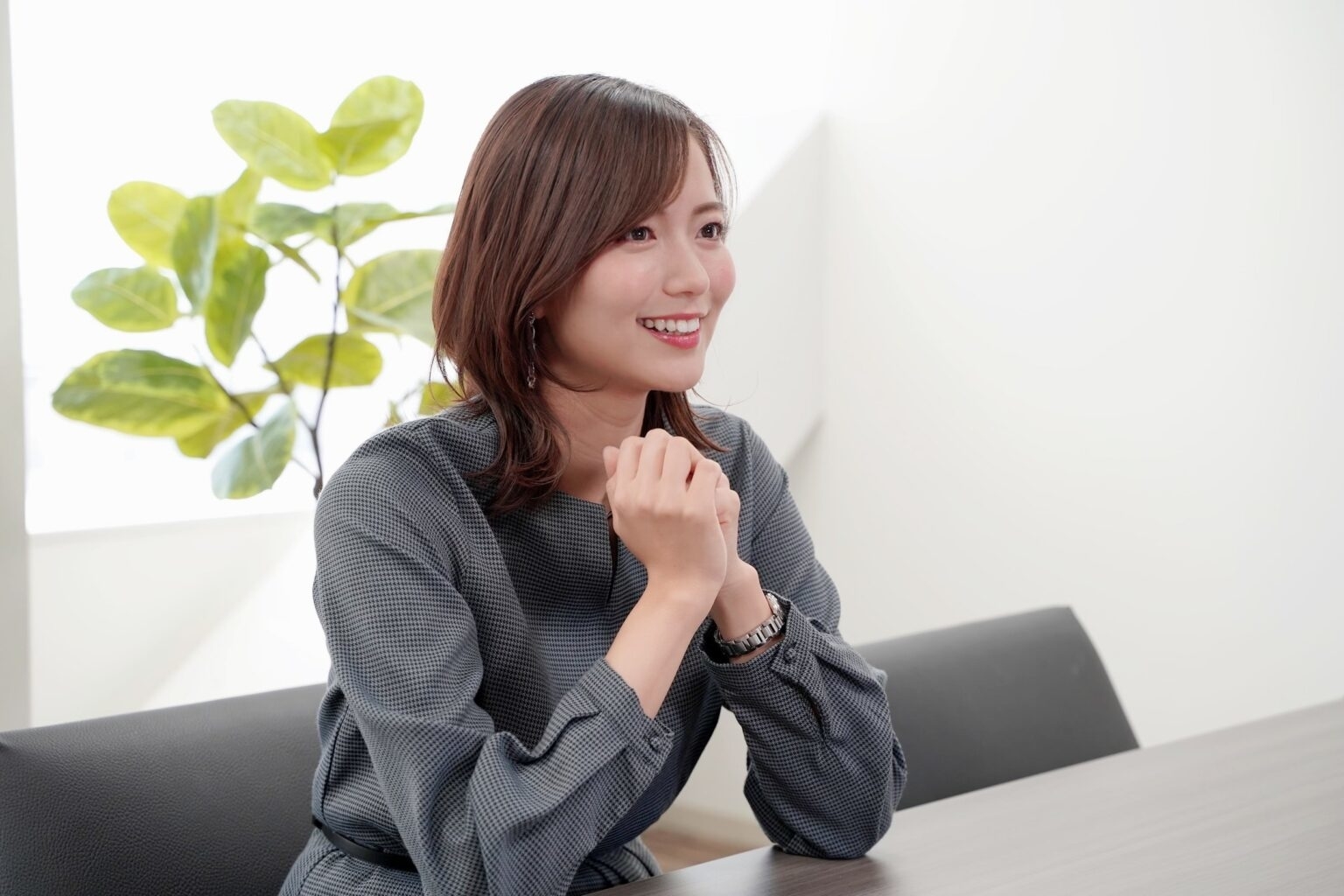 女性が会議室で笑顔で座っている。彼女はグレーの長袖ドレスを着ている。背景には植物が見える。