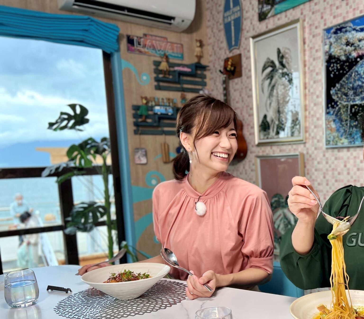 女性が明るく微笑んで食事をしているテーブル、一方にいる人がフォークで麺を持ち上げています。背景には海の景色が見えます。