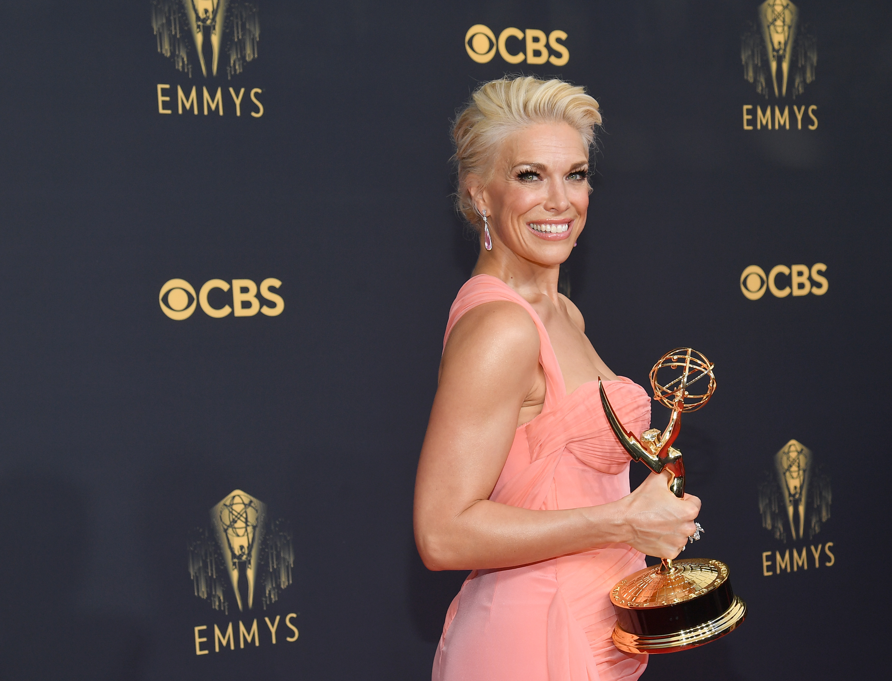 Hannah Waddingham, smiling and holding an Emmy award, poses on the red carpet at the Emmys. She is wearing an elegant sleeveless dress