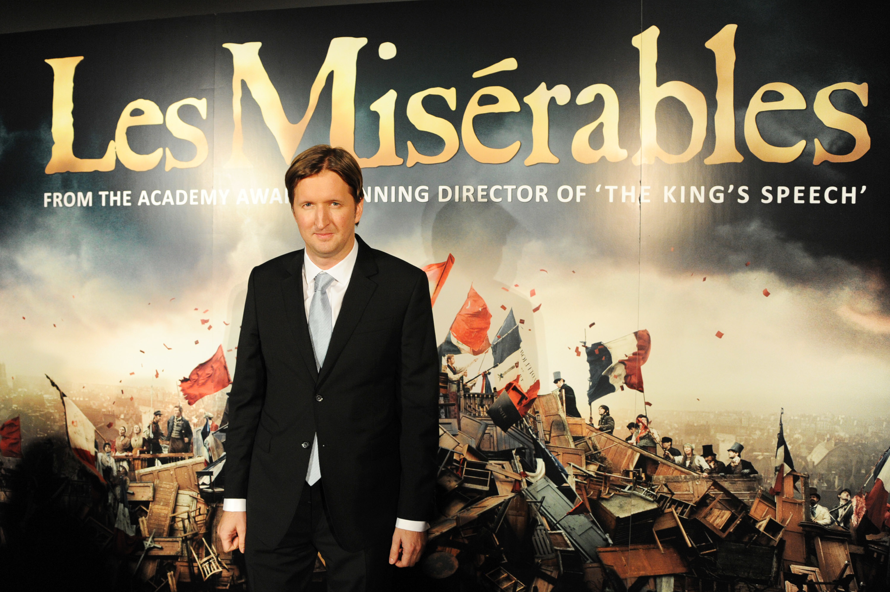 Tom Hooper on the red carpet at the premiere of &quot;Les Misérables,&quot; wearing a dark suit and tie, against a backdrop featuring the film&#x27;s title and a scene of the barricades