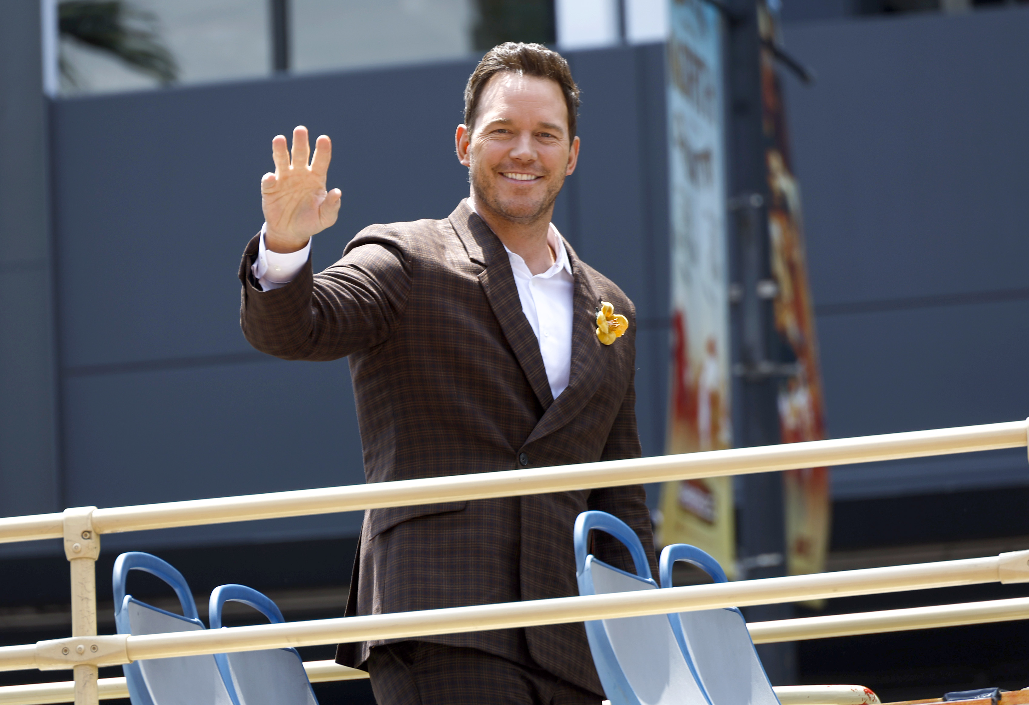 Chris Pratt waves while standing on an open-top bus, wearing a checkered suit and a pocket square