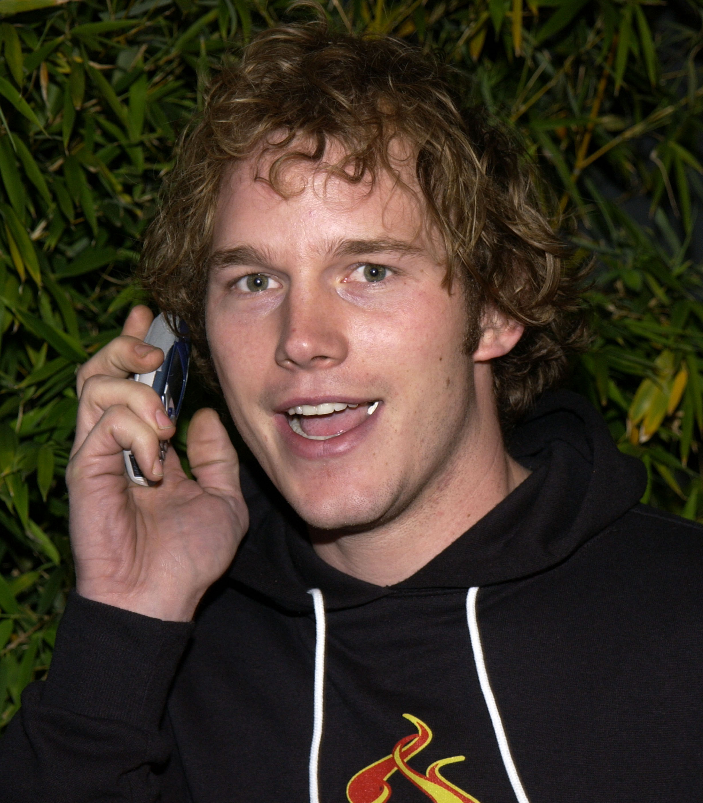 Chris Pratt smiles while speaking on a cellphone, wearing a hoodie with a flame design