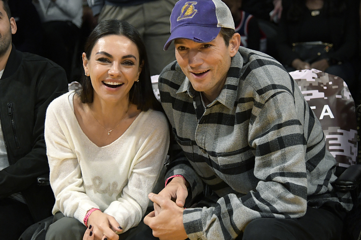 Mila Kunis and Ashton Kutcher sitting courtside at a basketball game, smiling and holding hands. Mila is wearing a white top, Ashton a plaid shirt and baseball cap