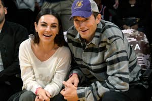 Mila Kunis and Ashton Kutcher sitting courtside at a basketball game, smiling and holding hands. Mila is wearing a white top, Ashton a plaid shirt and baseball cap