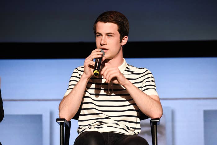 Dylan Minnette speaks into a microphone while seated on a stage, wearing a striped shirt