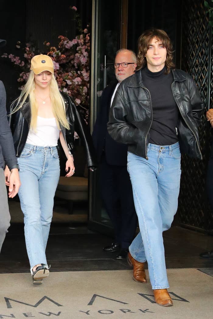 Anya Taylor-Joy and Malcolm McRae in casual outfits with leather jackets and jeans, walking out of a building
