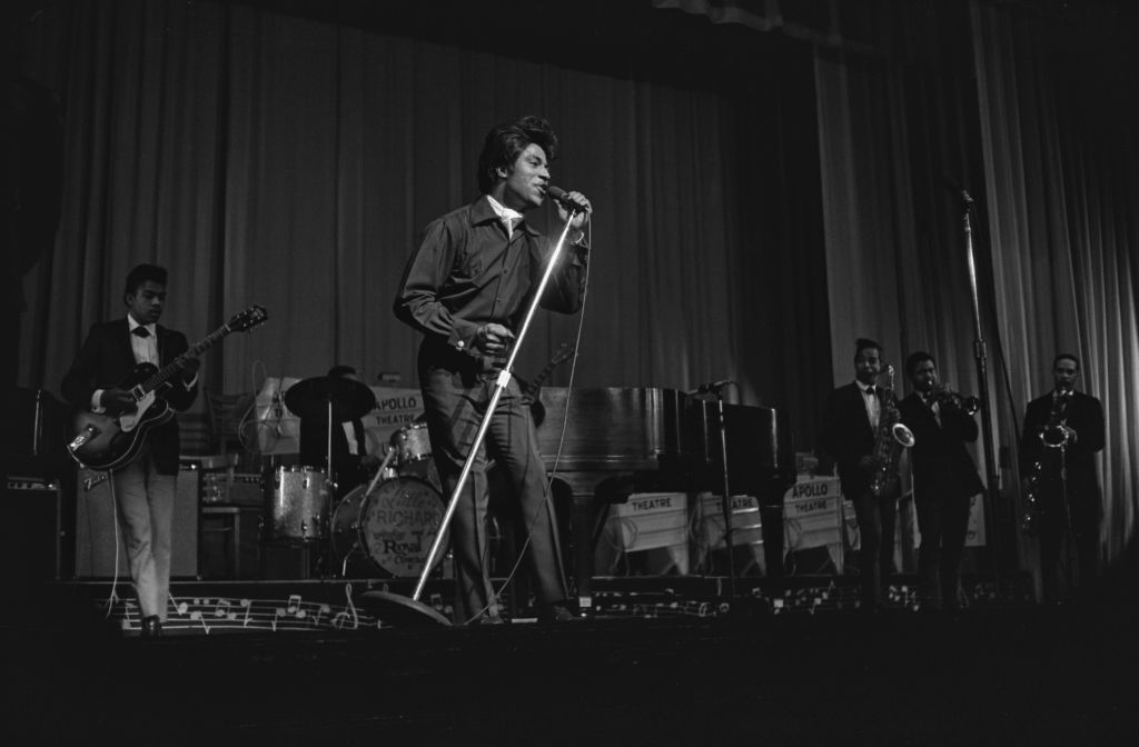 Little Richard, performs on stage with his band, singing into a microphone. Musicians, including Jim Hendrix, playing various instruments stand in the background
