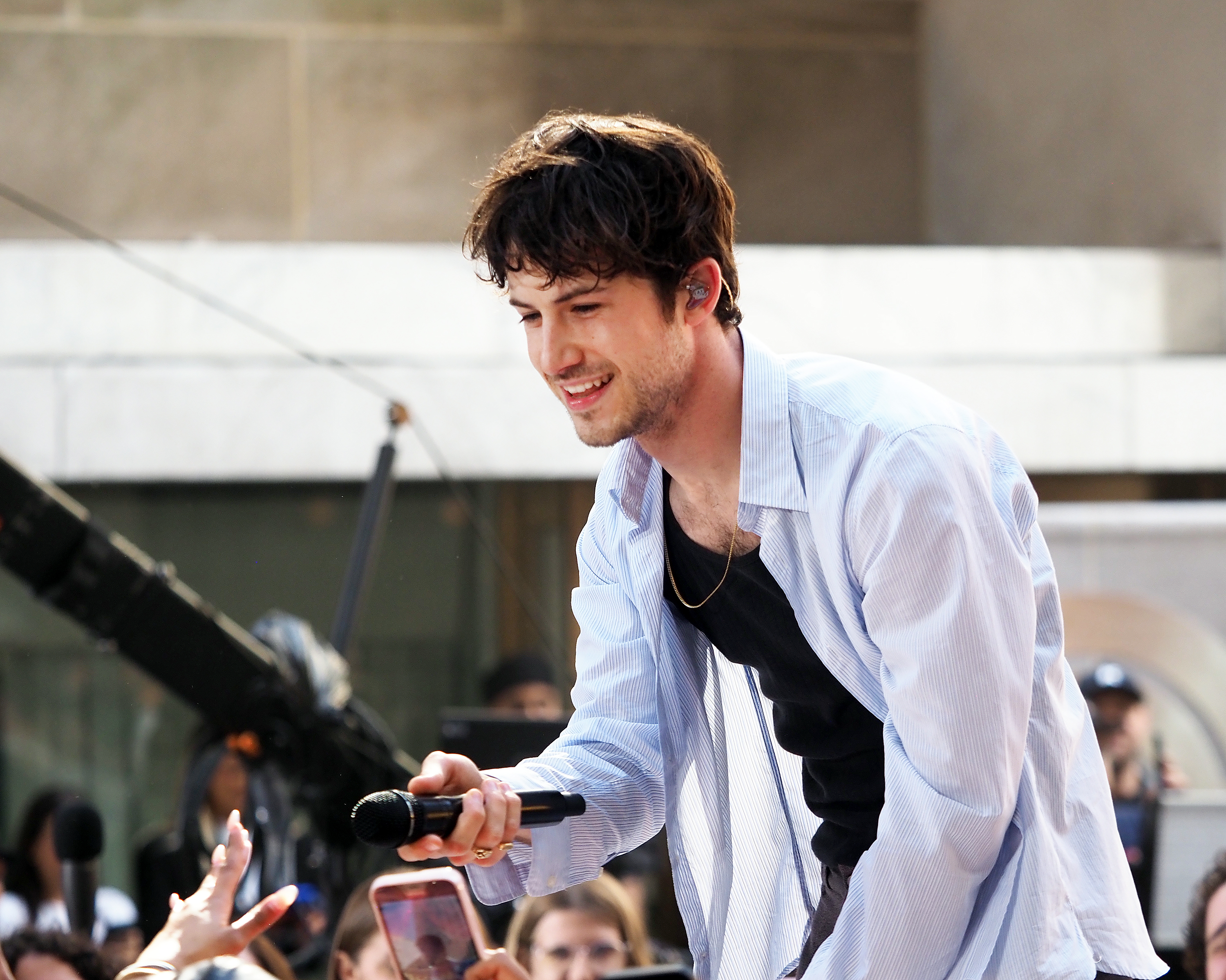 Dylan Minnette, wearing a light shirt over a tank top, smiles as he interacts with fans during a live performance