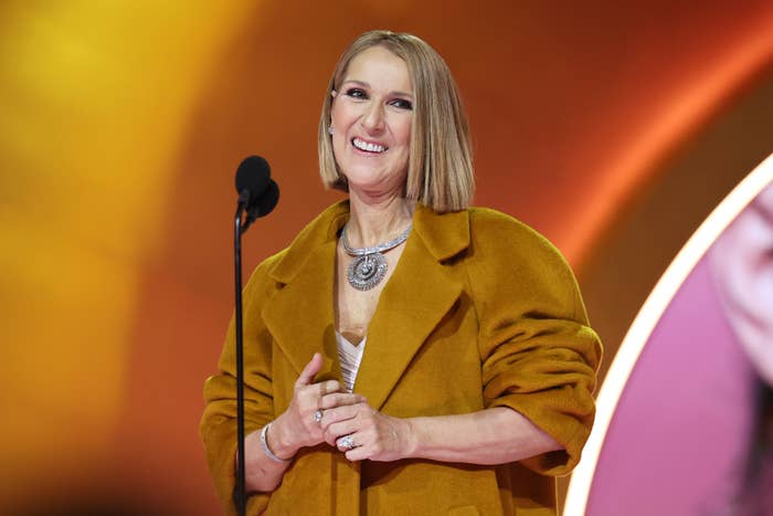 Celine Dion, smiling, stands at a microphone wearing an oversized coat and a statement necklace