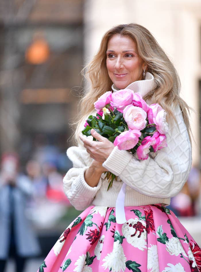 Celine Dion holding a bouquet of flowers, wearing a cozy turtleneck sweater and a floral-patterned skirt, smiling outdoors