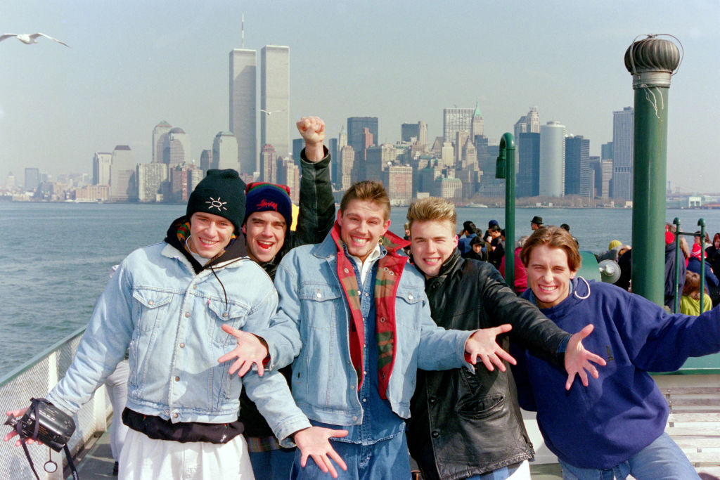 Five members of Take That posing energetically with New York City skyline in the background, all dressed in casual jackets