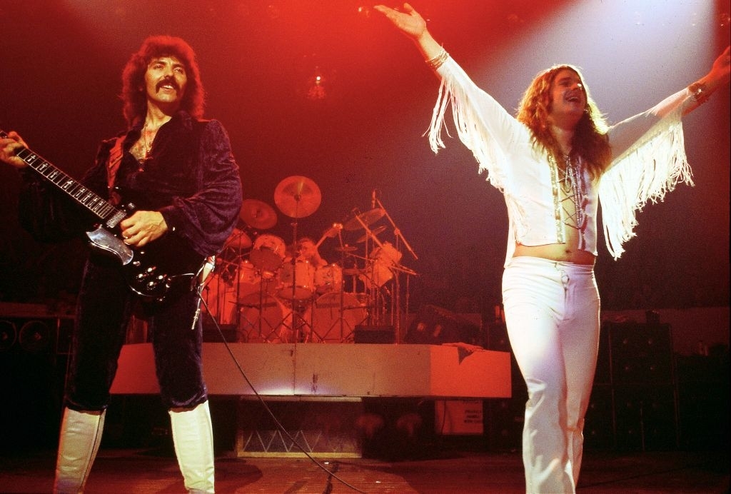 Tony Iommi playing guitar in a velvet suit and Ozzy Osbourne performing with arms outstretched in a fringe jacket on stage, with a drummer in the background