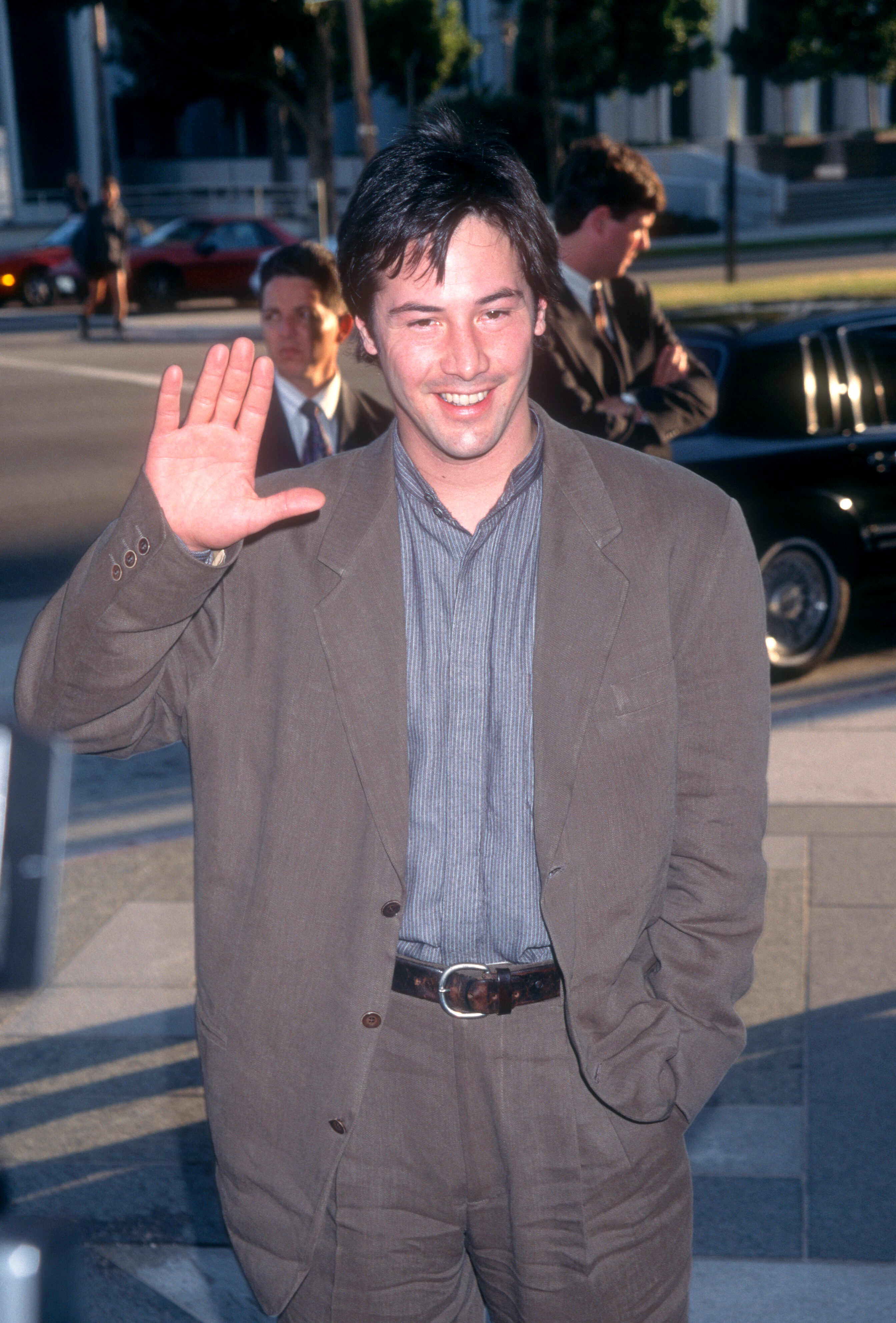 Keanu Reeves waves while wearing a casual suit, standing outdoors during an event. Two other individuals are visible in the background