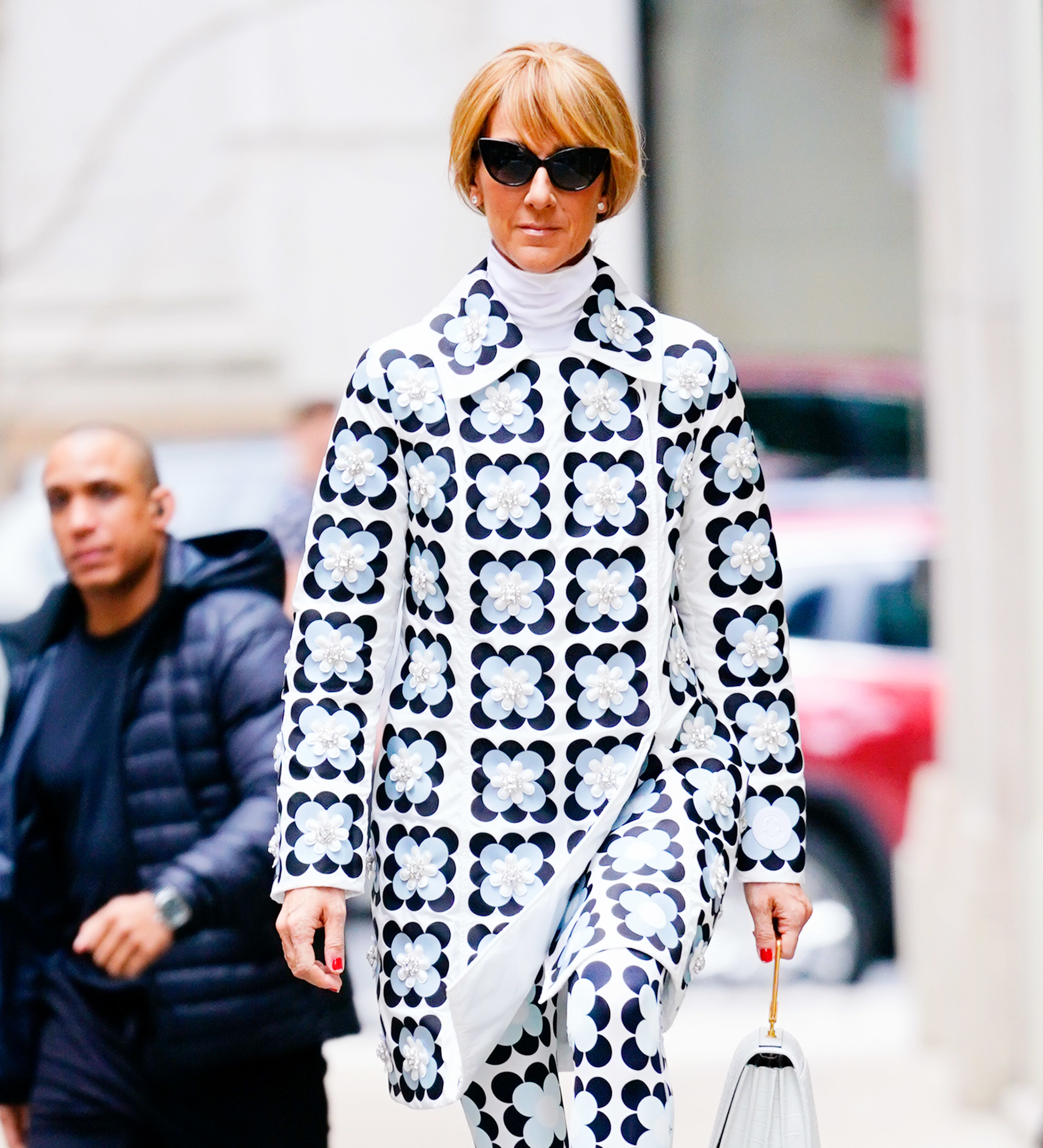 Celine Dion walking outdoors in a polka-dot patterned outfit with a matching coat and sunglasses, holding a purse