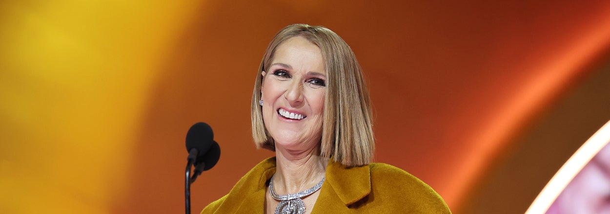 Celine Dion, smiling, stands at a microphone wearing an oversized coat and a statement necklace