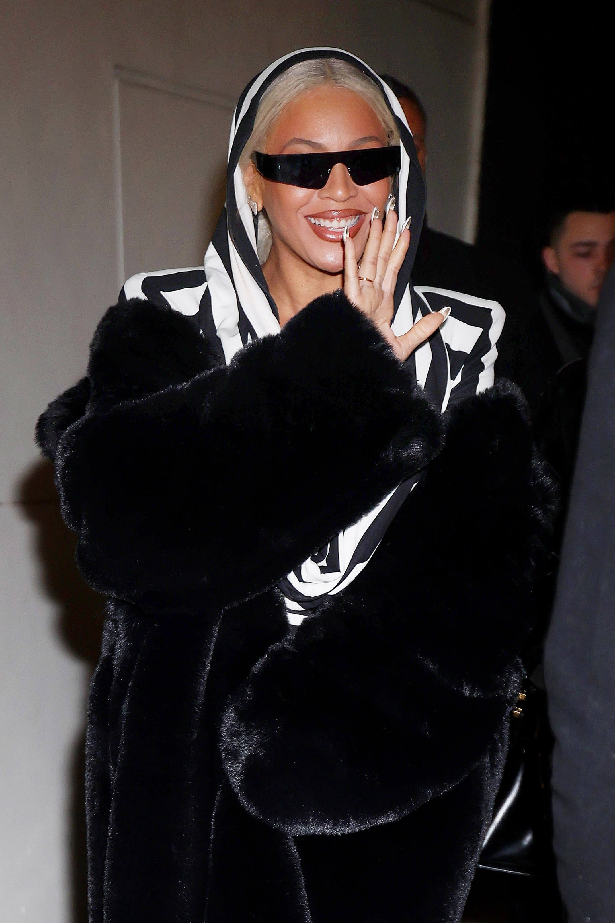 Beyoncé smiles and waves while wearing a fur coat, sunglasses, and -hooded outfit
